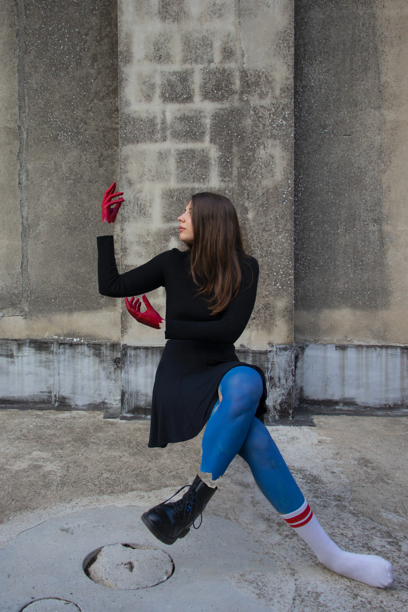 A portrait: a woman in black with red gloves and blue tights, against a concrete wall.