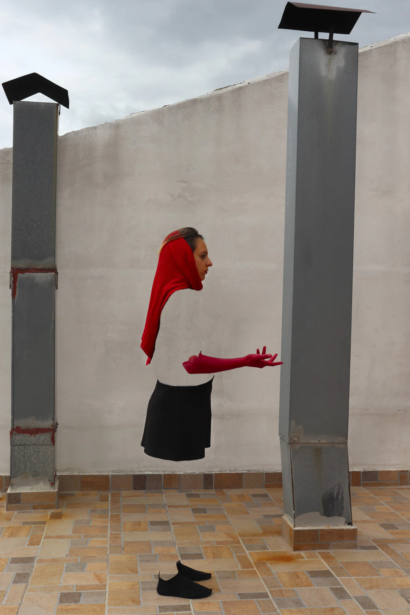 A portrait: a woman with a red hood and red gloves, arms forward, between two chimneys on a roof.