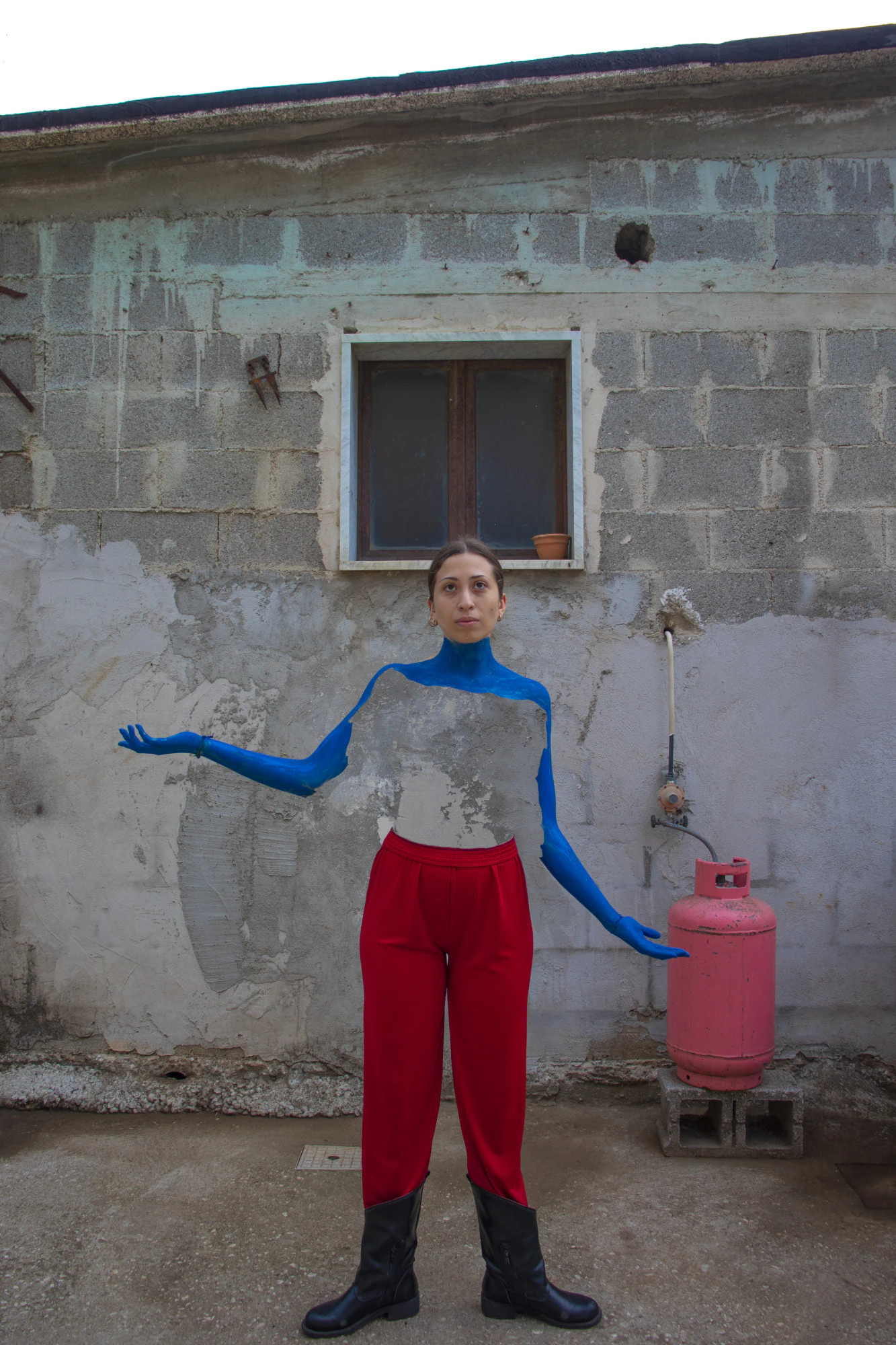 A portrait: a woman with blue sleeves and red trousers, beside a gas cylinder.