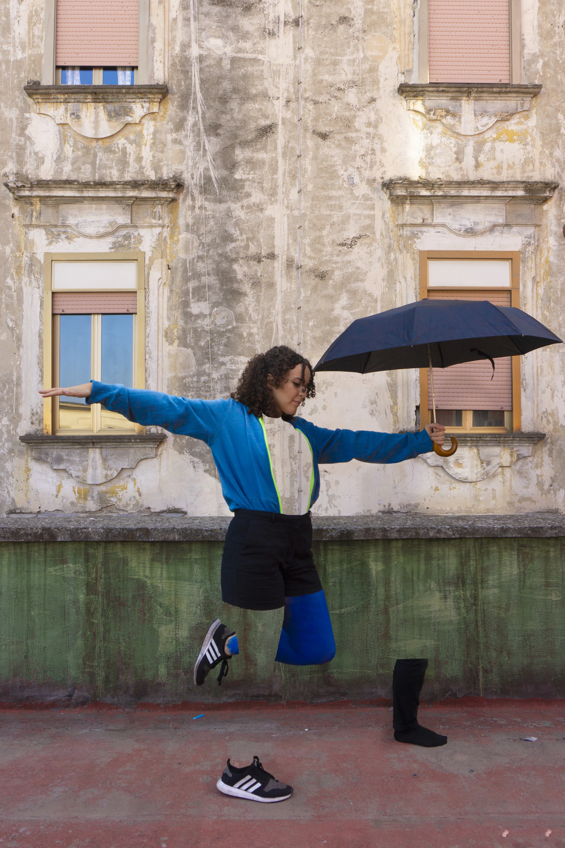 A portrait: a woman with an umbrella and blue sleeves, leaping in front of a building.