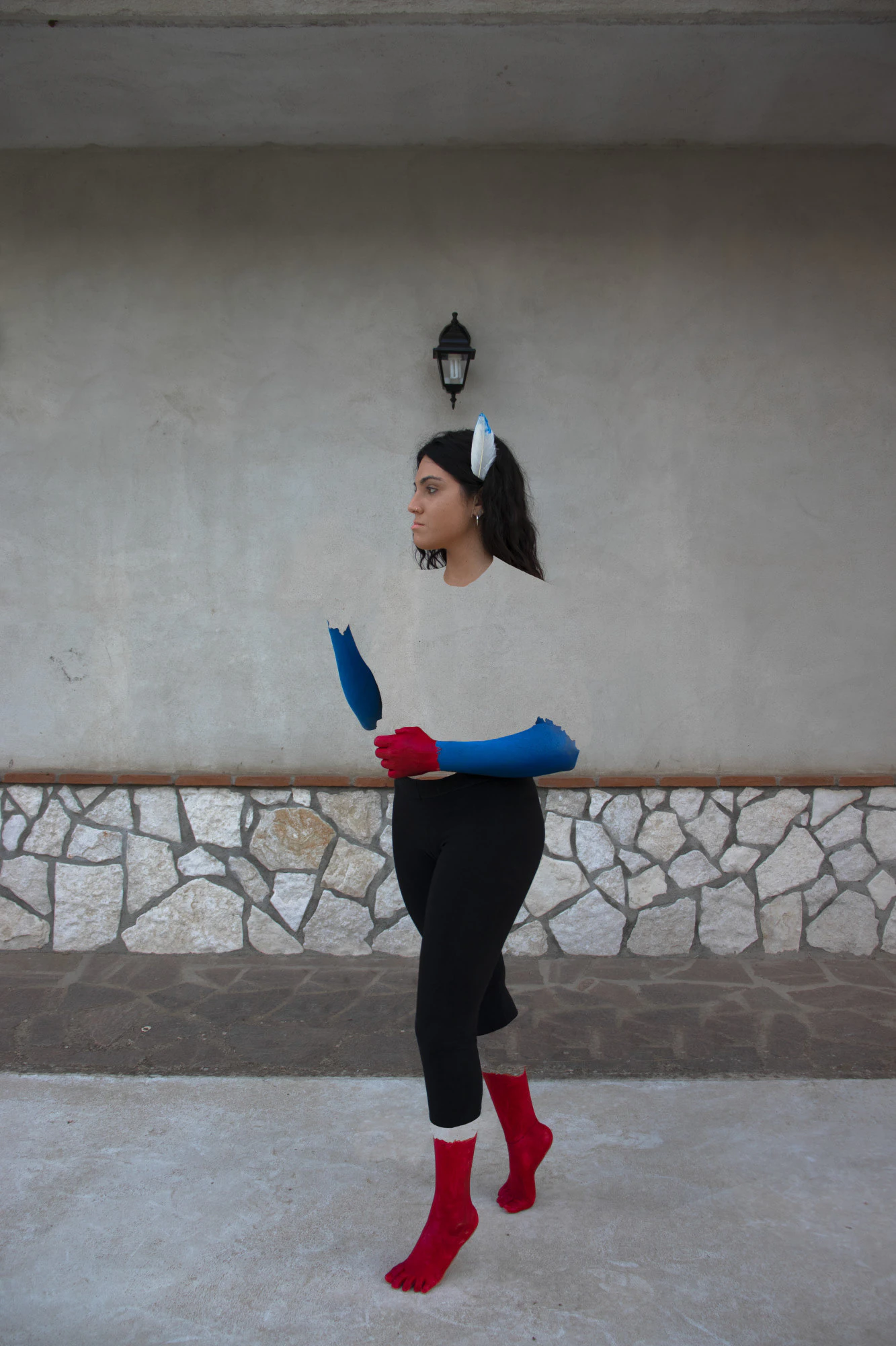 A portrait: a woman with blue and red gloves and red socks, dancing in front of a wall.